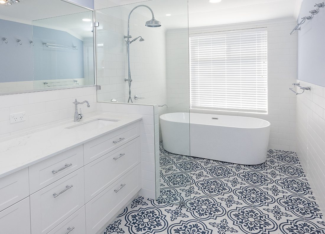 This large bathroom renovation features a freestanding bathtub, dark blue and white ornamental floor tiling, and two sinks below a large rectangular mirror