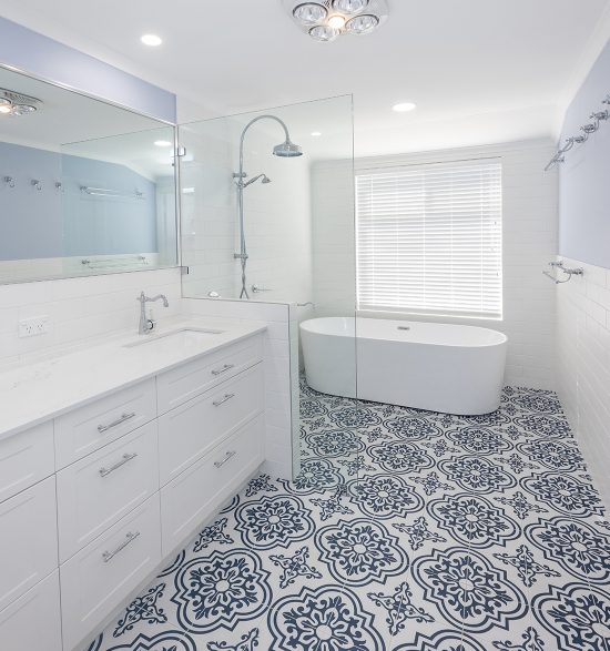 This large bathroom renovation features a freestanding bathtub, dark blue and white ornamental floor tiling, and two sinks below a large rectangular mirror