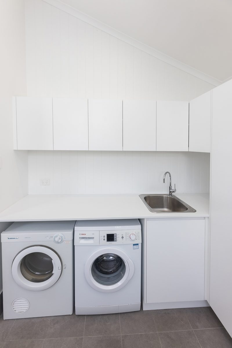 A laundry area with a drop-in sink, chrome tapware, a large countertop with a washing and drying machine stored underneath, and storage cupboards