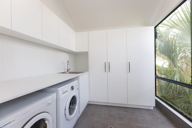 A laundry renovation that features new white storage cupboards, a washer and dryer space under the sink countertop, and large windows with a view of outside greenery