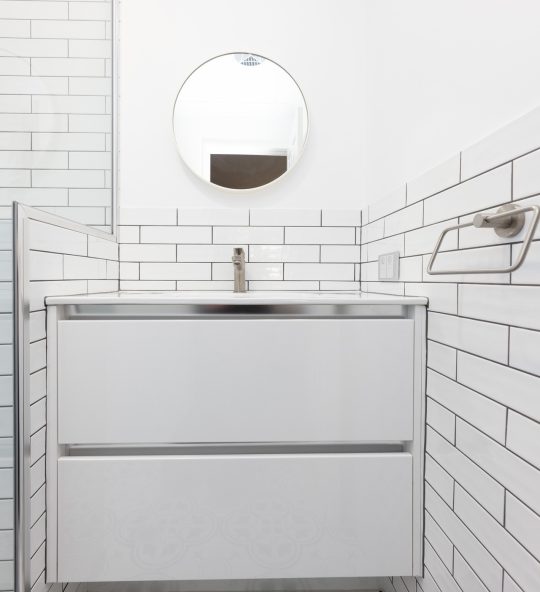 White vanity with a small circular mirror above and white tiles