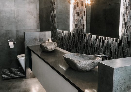 A modern bathroom featuring black and gray tiling on the double vanity backsplash and floor, and free-form shaped stone sink basins