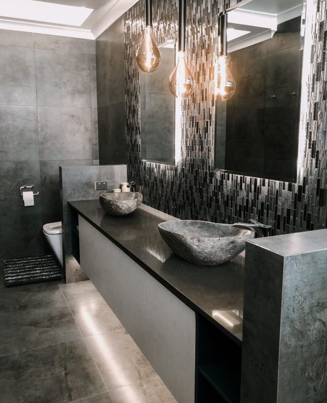 A modern bathroom featuring black and gray tiling on the double vanity backsplash and floor, and free-form shaped stone sink basins