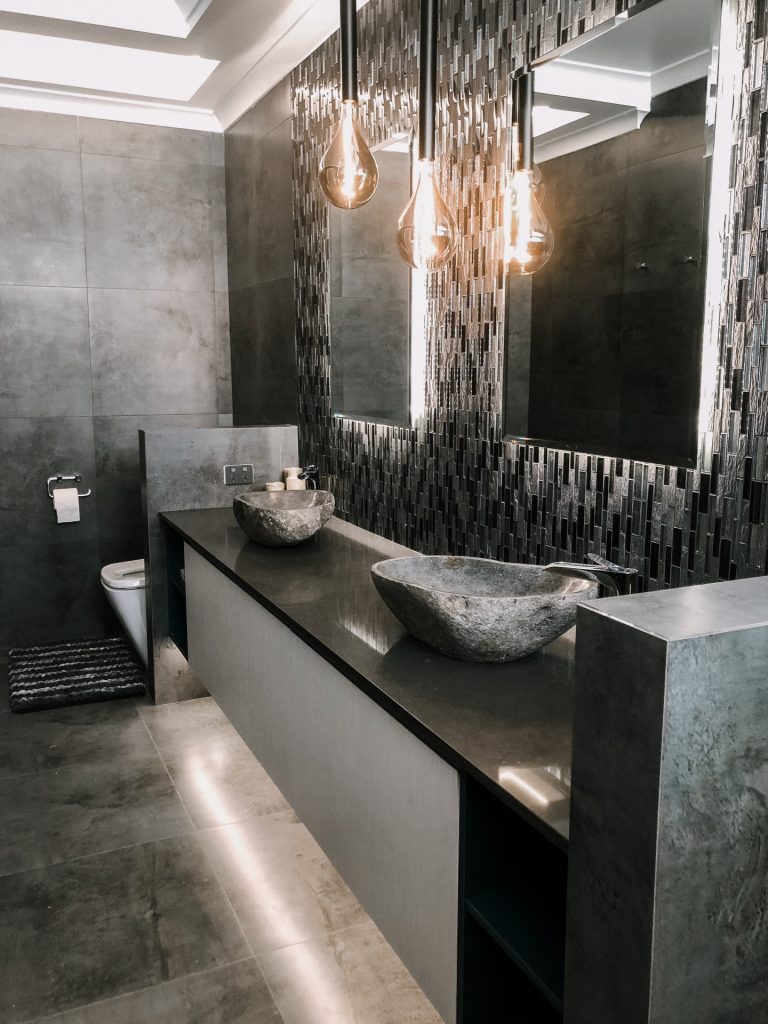 A modern bathroom featuring black and gray tiling on the double vanity backsplash and floor, and free-form shaped stone sink basins